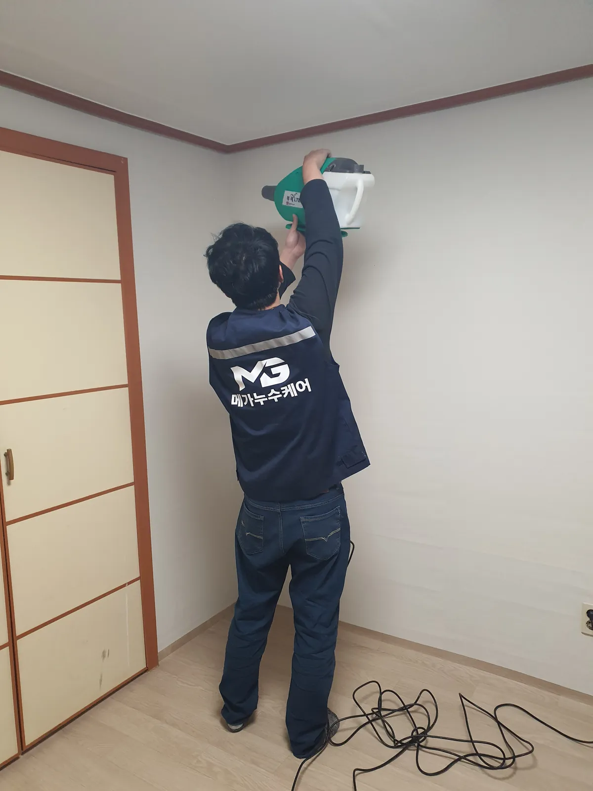 새 흰 벽지로 마감된 방에서 작업자가 초록색 전기 분무기를 머리 위로 들어 천장 쪽으로 뿌리는 뒷모습