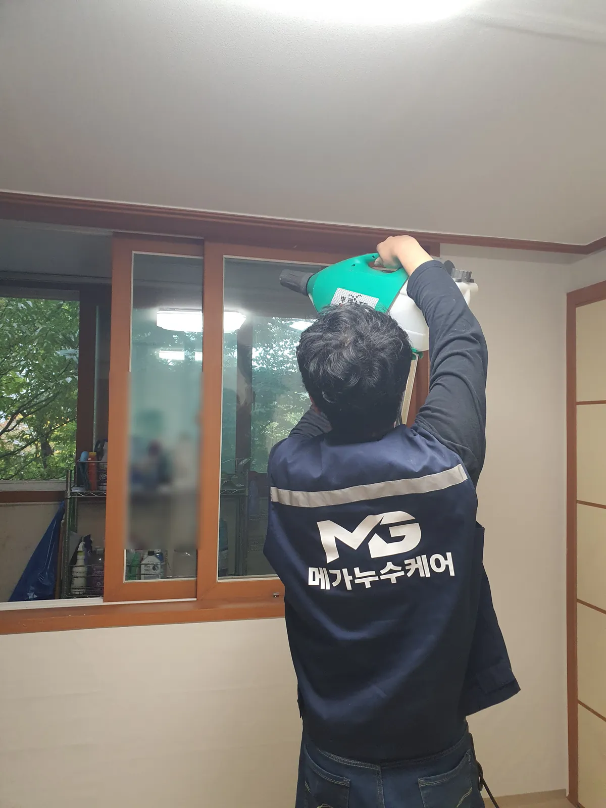 나무 창틀 위쪽 벽을 향해 작업자가 초록색 전기 분무기를 들어 뿌리는 뒷모습